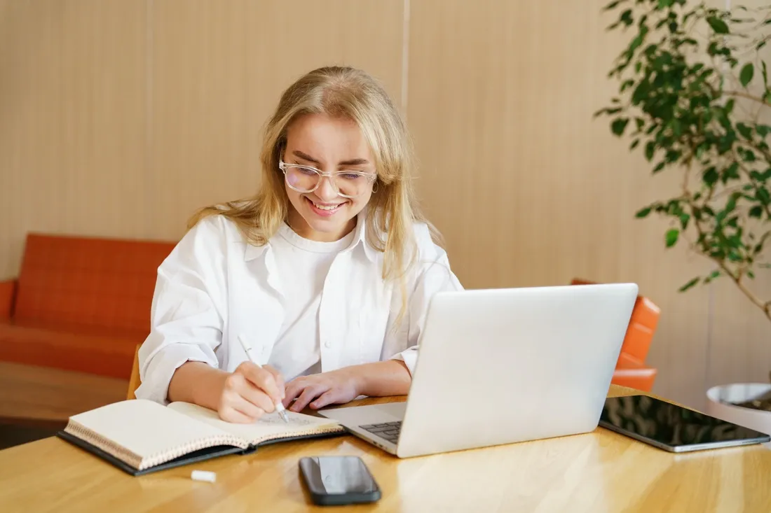 Femme souriante entrain d'écrire sur un cahier et avec son ordinateur portable devant elle