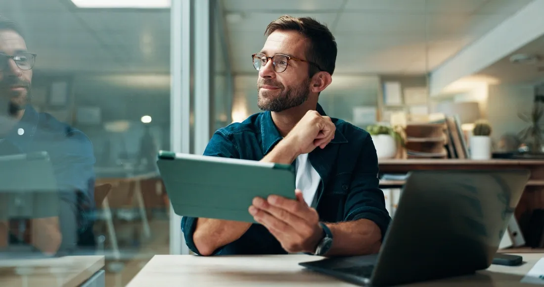 Homme souriant travaillant sur une tablette numérique dans un bureau moderne, avec un ordinateur portable posé sur la table