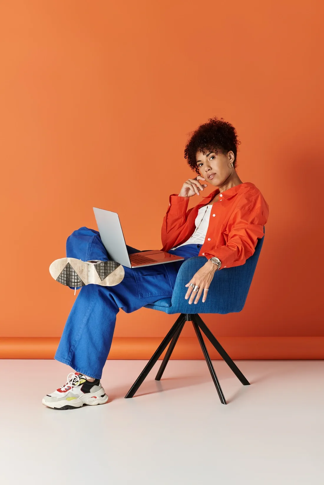 Jeune femme assise sur une chaise bleue, habillée en tenue décontractée colorée, utilisant un ordinateur portable devant un mur orange.