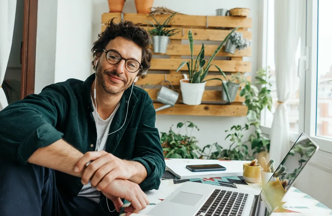 Un homme souriant avec des écouteurs filaires et assis à son bureau avec son ordinateur portable