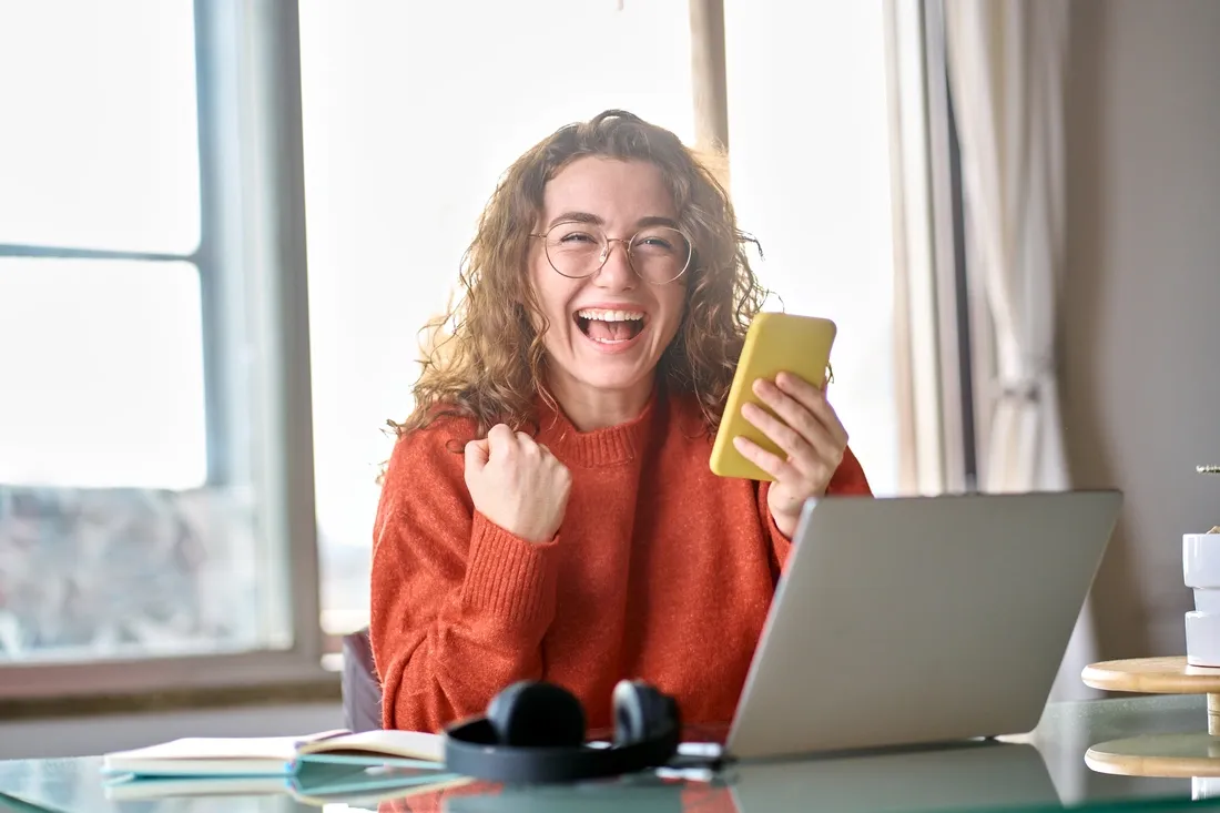 Personne célébrant une bonne nouvelle au téléphone, assise devant un ordinateur portable