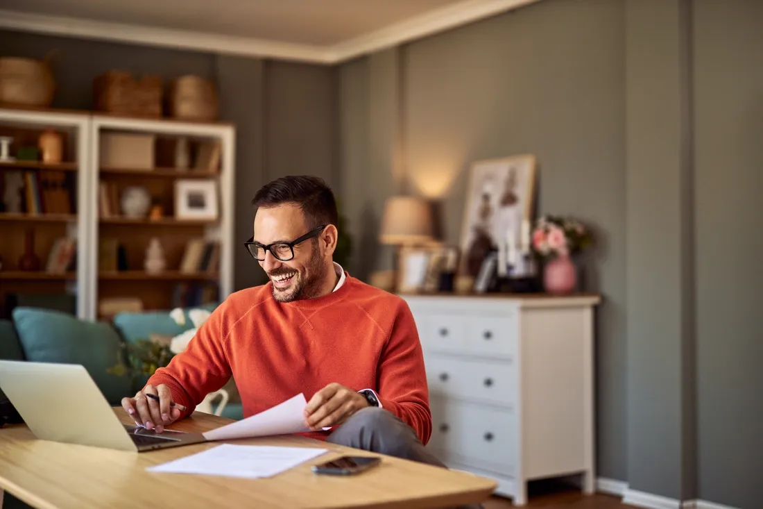 Homme avec un pull orange chez lui entrain d'utiliser son ordinateur portable sur une table à manger