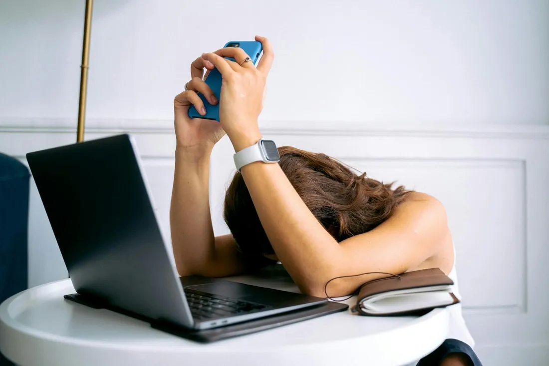 Femme assise à une table devant un ordinateur portable, la tête posée sur ses bras et tenant son téléphone avec lassitude