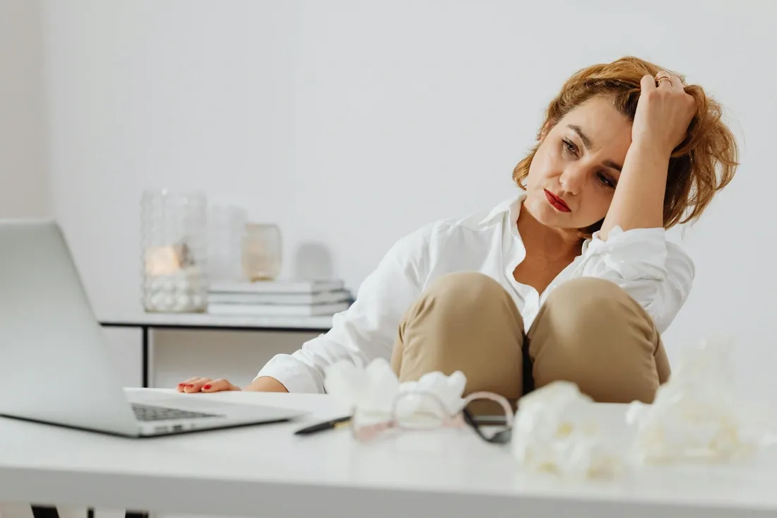 Femme assise devant un ordinateur portable, l’air fatigué et la tête appuyée sur sa main, entourée de papiers froissés